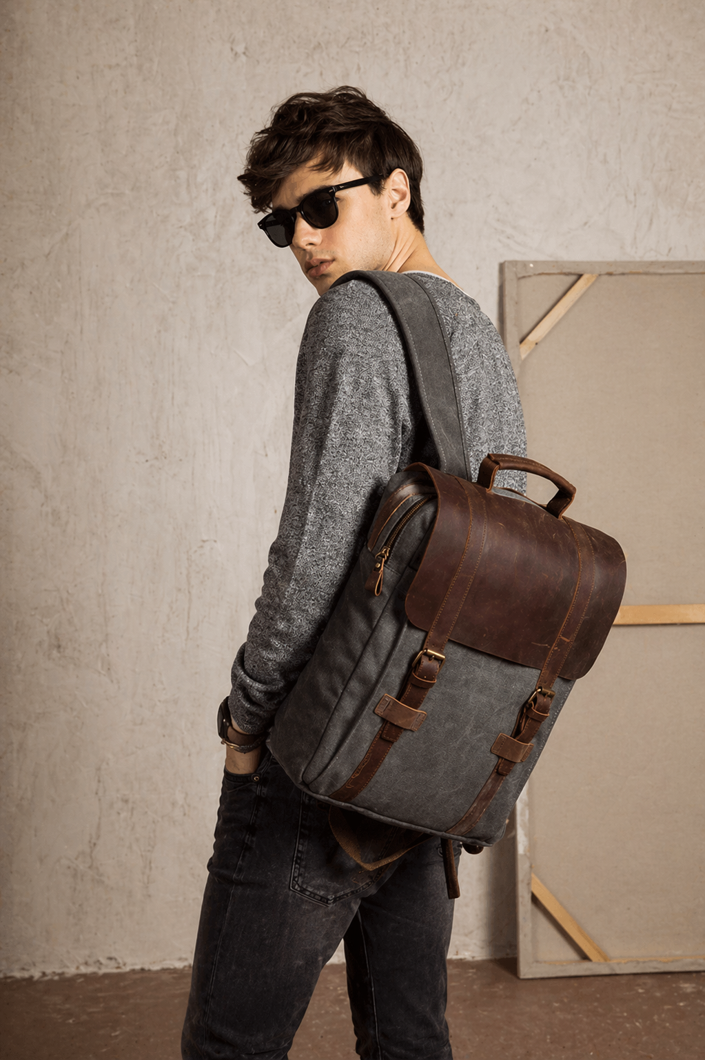 Person wearing a gray backpack with brown leather straps against a plain wall.