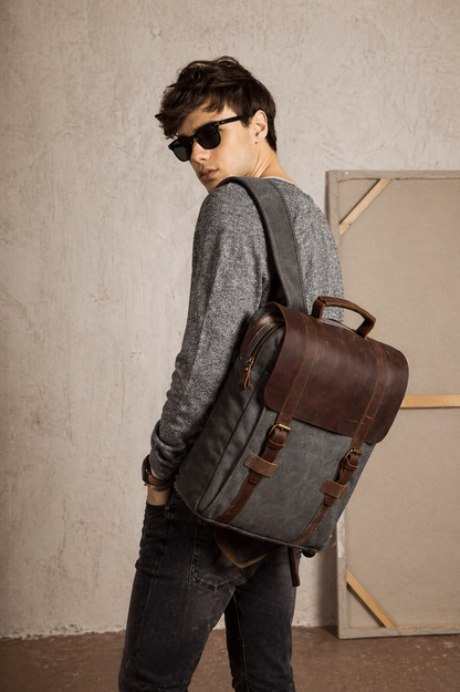 Person wearing a gray backpack with brown leather straps against a plain wall.