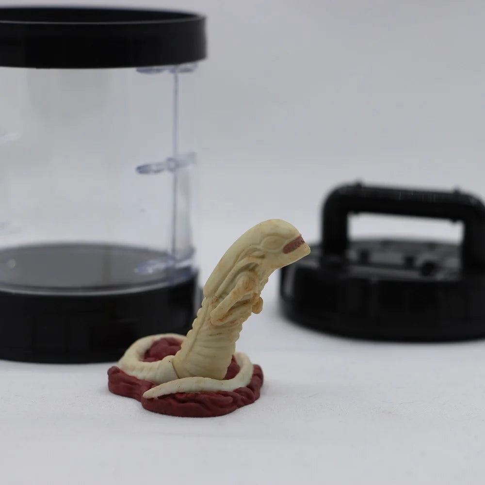 Small figurine of a creature with a long tongue on a white surface, with a clear container and black lid in the background.