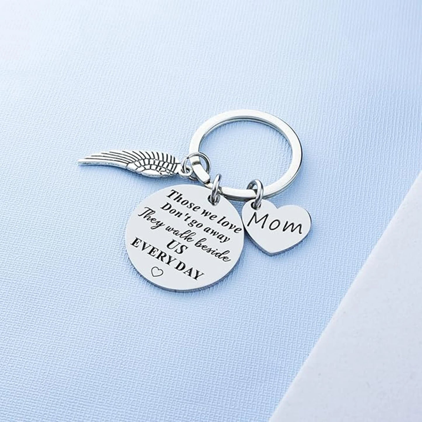 Silver keychain with angel wing, heart-shaped 'Mom' tag, and engraved message on a light blue background.
