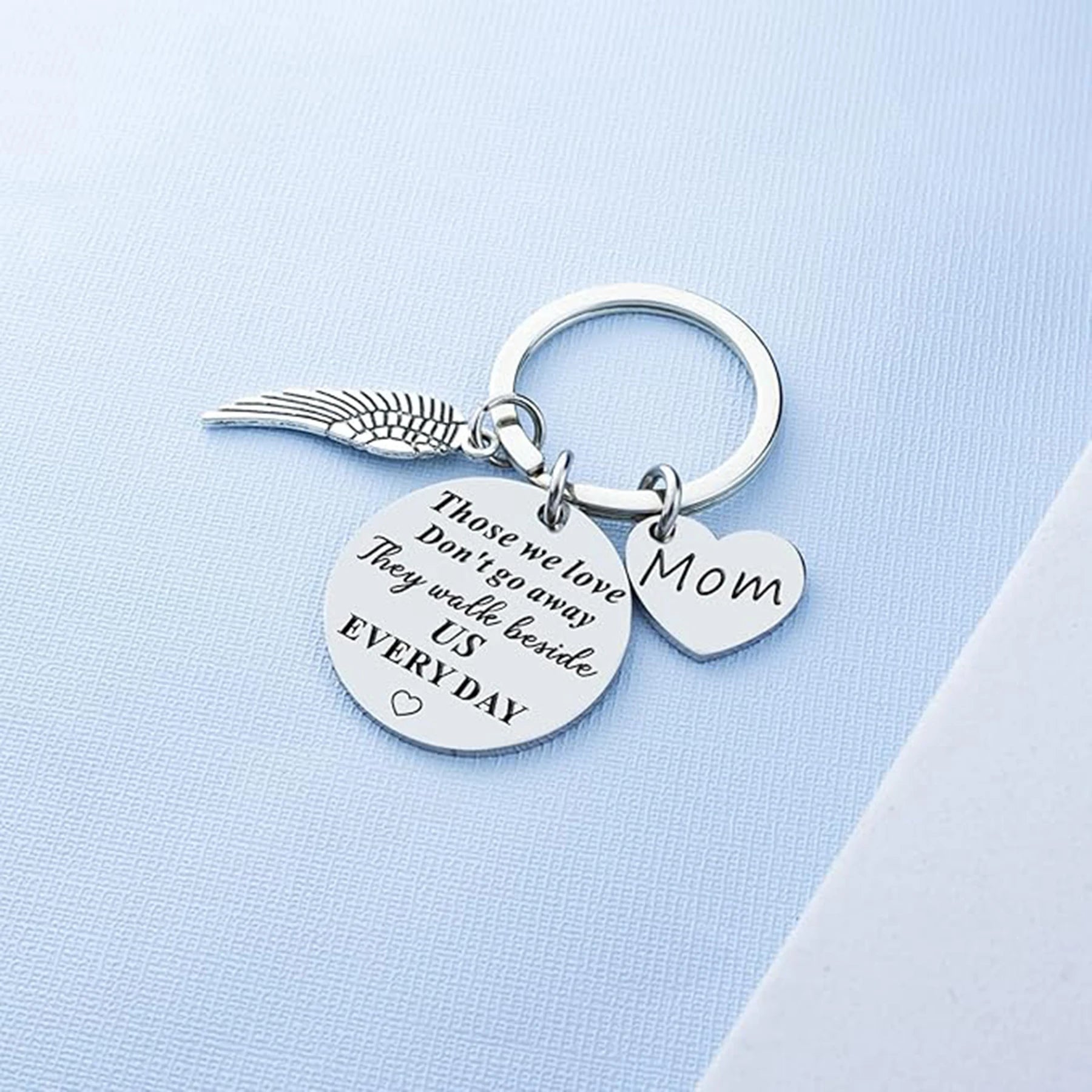 Silver keychain with angel wing, heart-shaped 'Mom' tag, and engraved message on a light blue background.