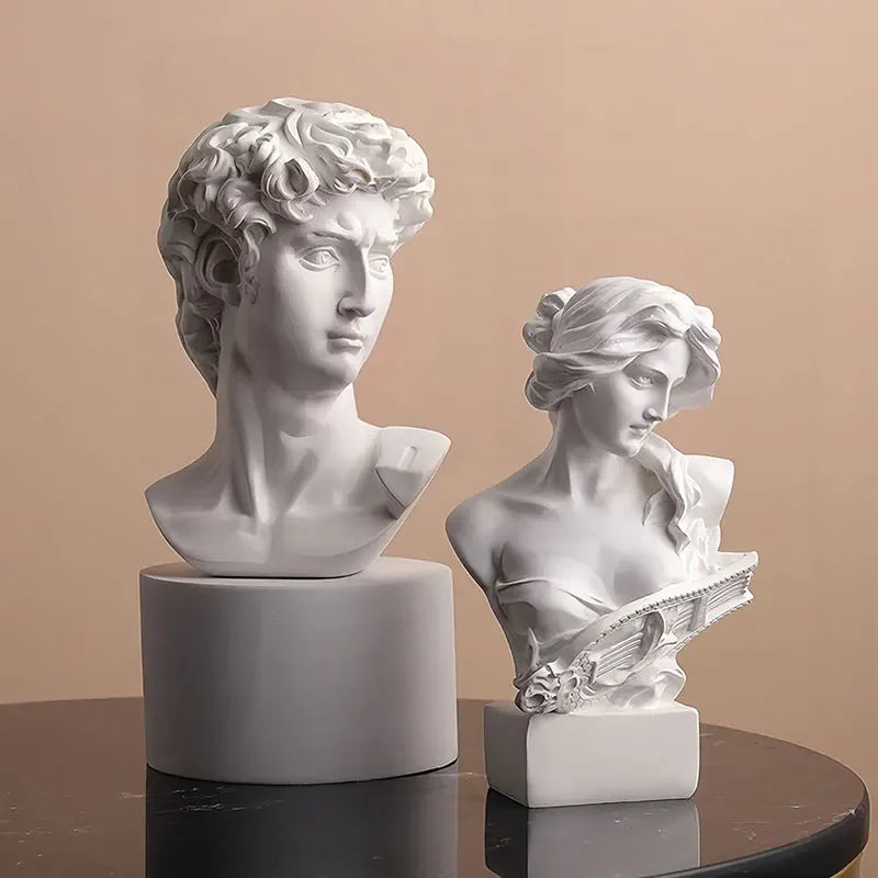 Two white marble busts of a man and a woman on a reflective surface with a beige background
