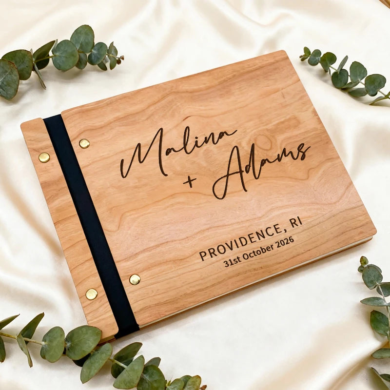 Wooden album with engraved names and location, surrounded by eucalyptus leaves on a white fabric background