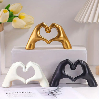 Decorative figurines of hands forming hearts in gold, white, and black on a surface with flowers and a lamp in the background.