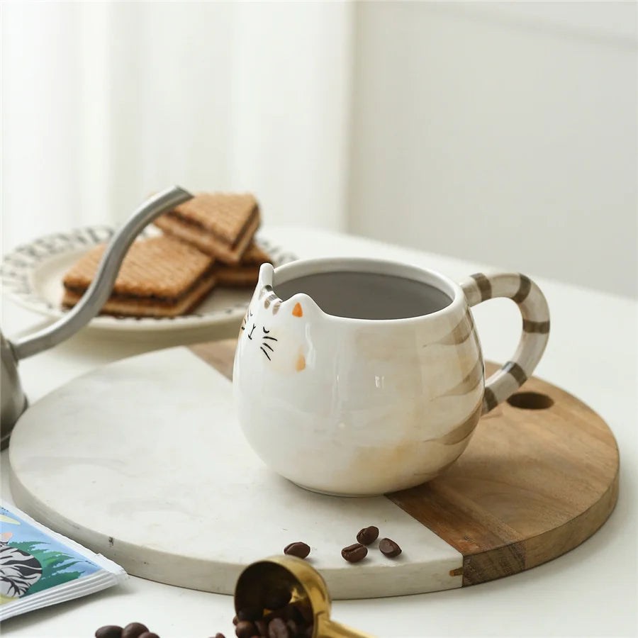 Cat-shaped ceramic mug on a wooden coaster with coffee beans and a plate of cookies in the background.