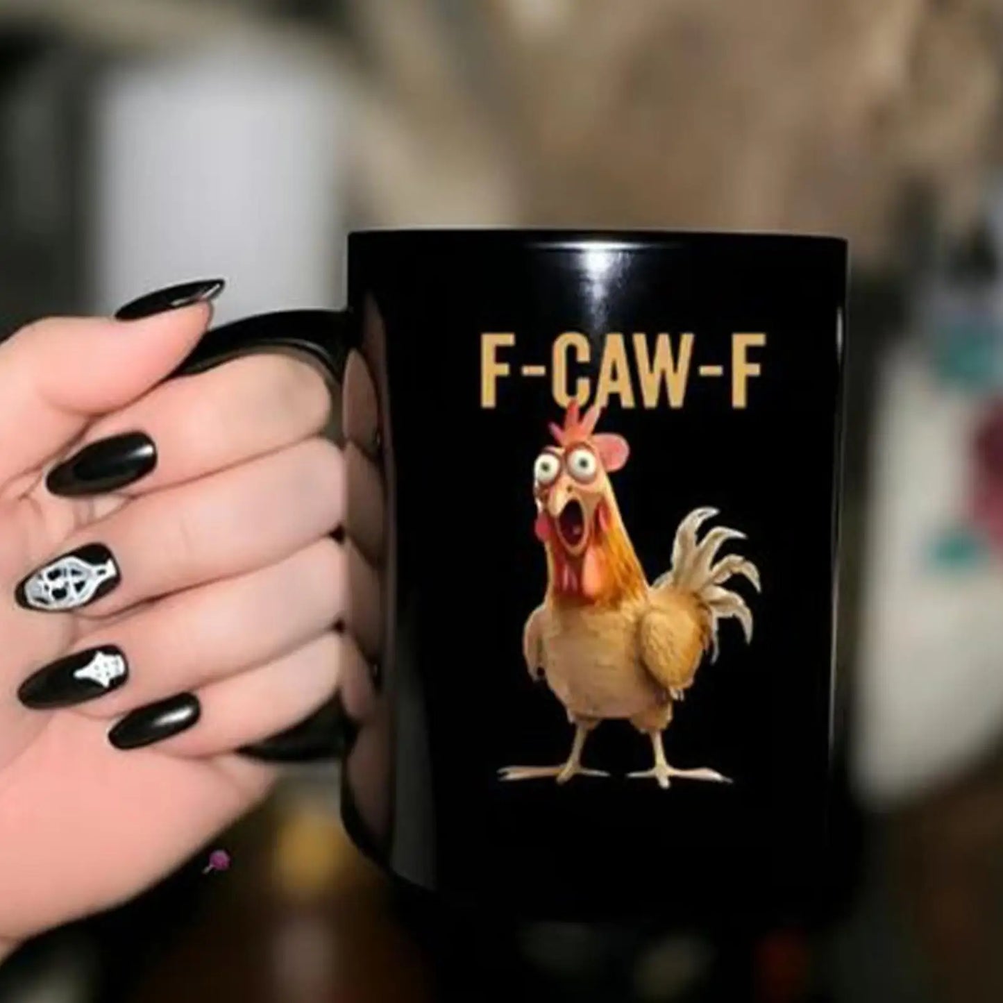 Black mug with a cartoon chicken and 'F-CAW-F' text held by a hand with black nail polish.