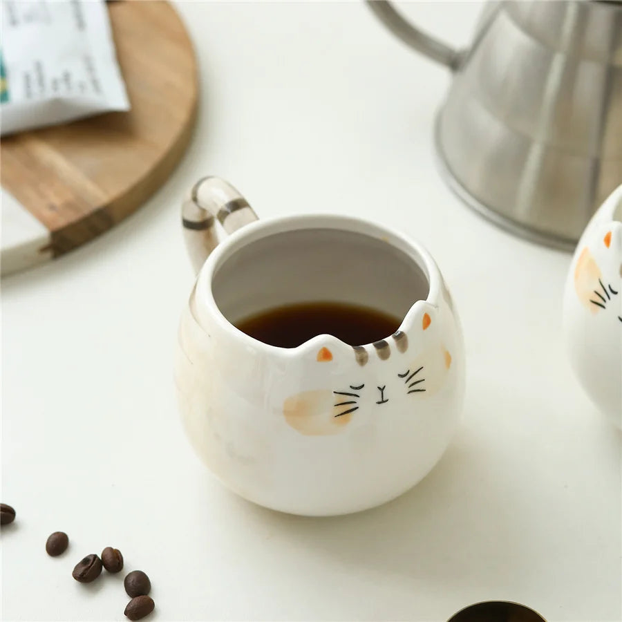 Cat-shaped ceramic mug with coffee on a white surface with coffee beans and a kettle.