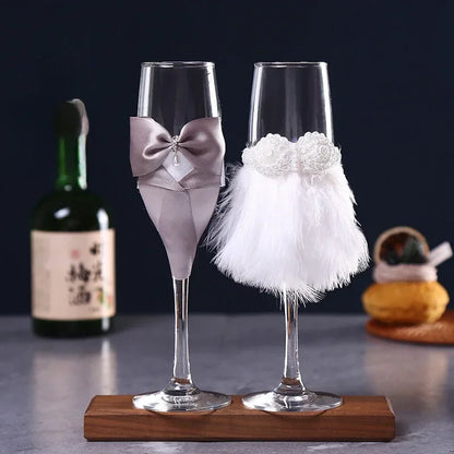 Two decorative champagne flutes with bows and feathers on a wooden stand, with a bottle of wine in the background.