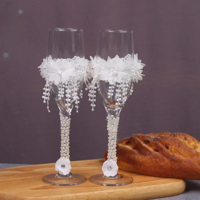Two decorative wine glasses with lace and beads on a wooden board with bread in the background.