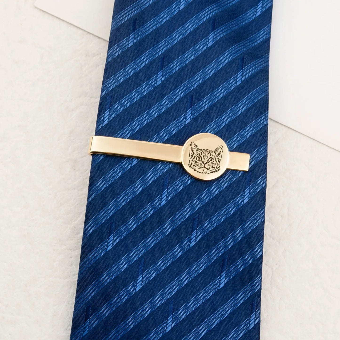 Gold tie clip with cat design on a blue striped tie against a white background