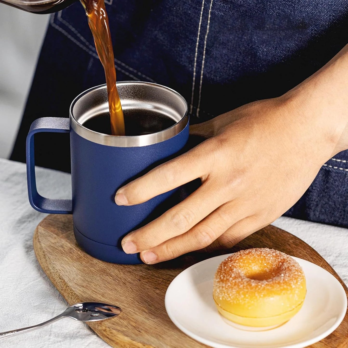 Person pouring coffee into a blue insulated mug with a donut on a wooden coaster.
