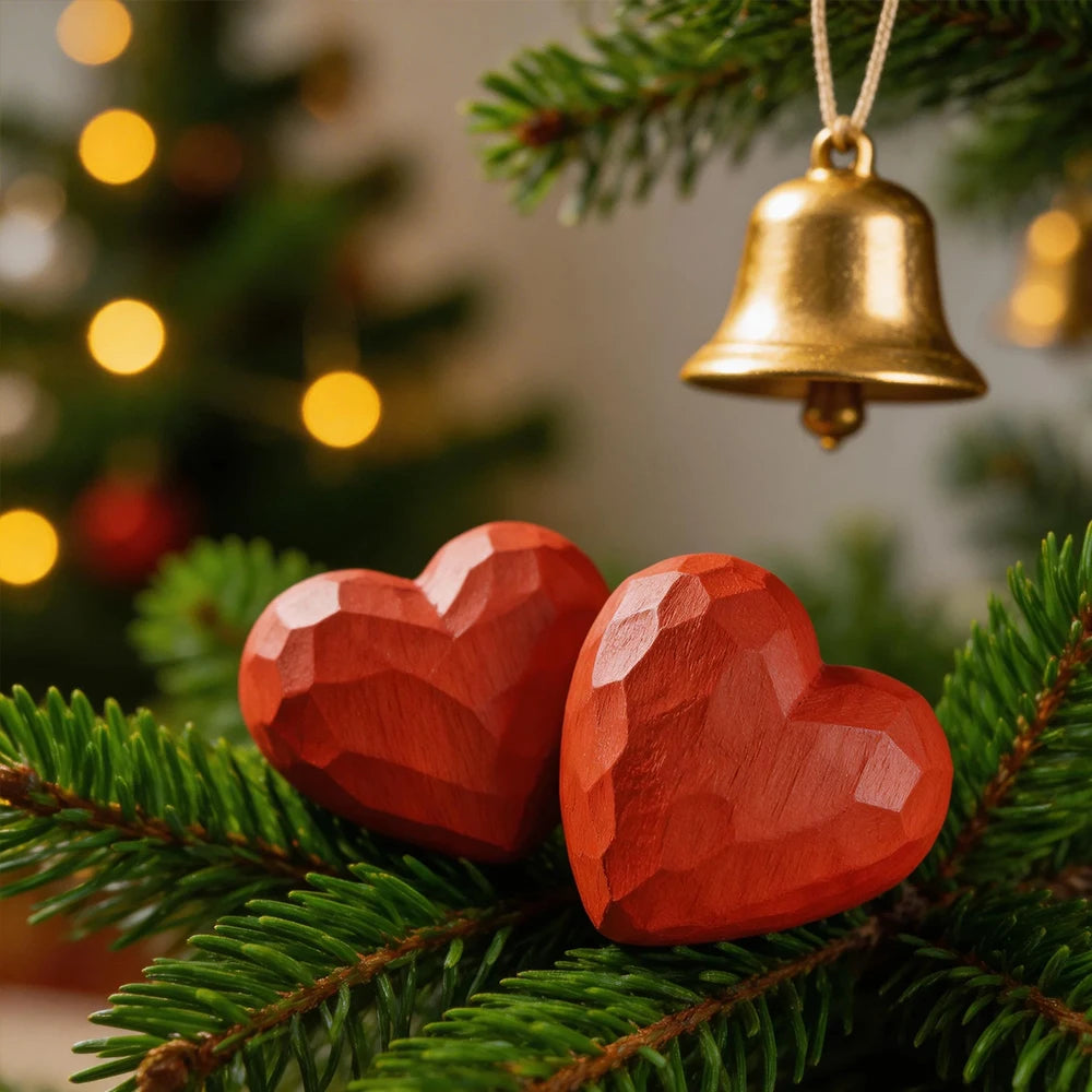 Red heart-shaped ornaments and a gold bell hanging on a Christmas tree branch with blurred lights in the background.