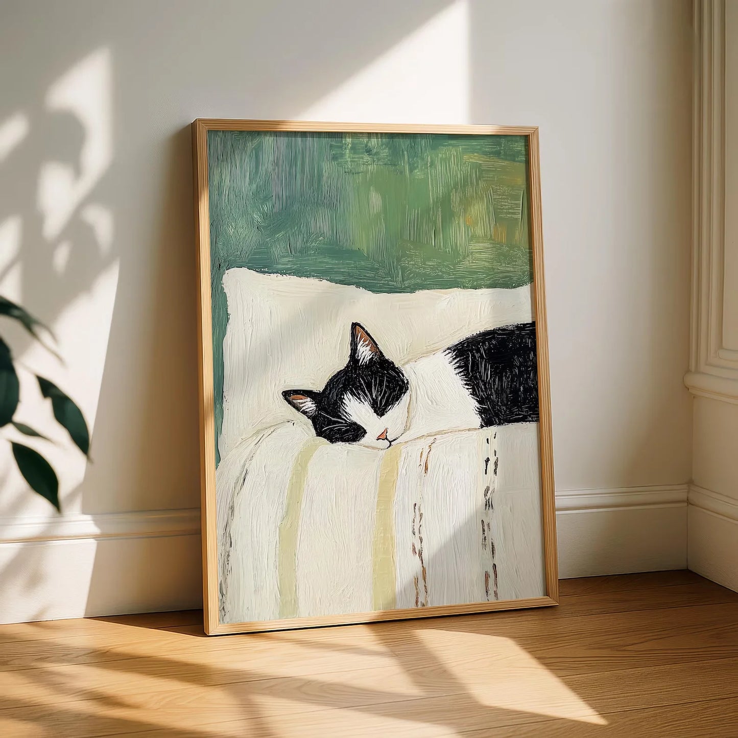Framed artwork of a black and white cat on a pillow with a green background, leaning against a white wall.