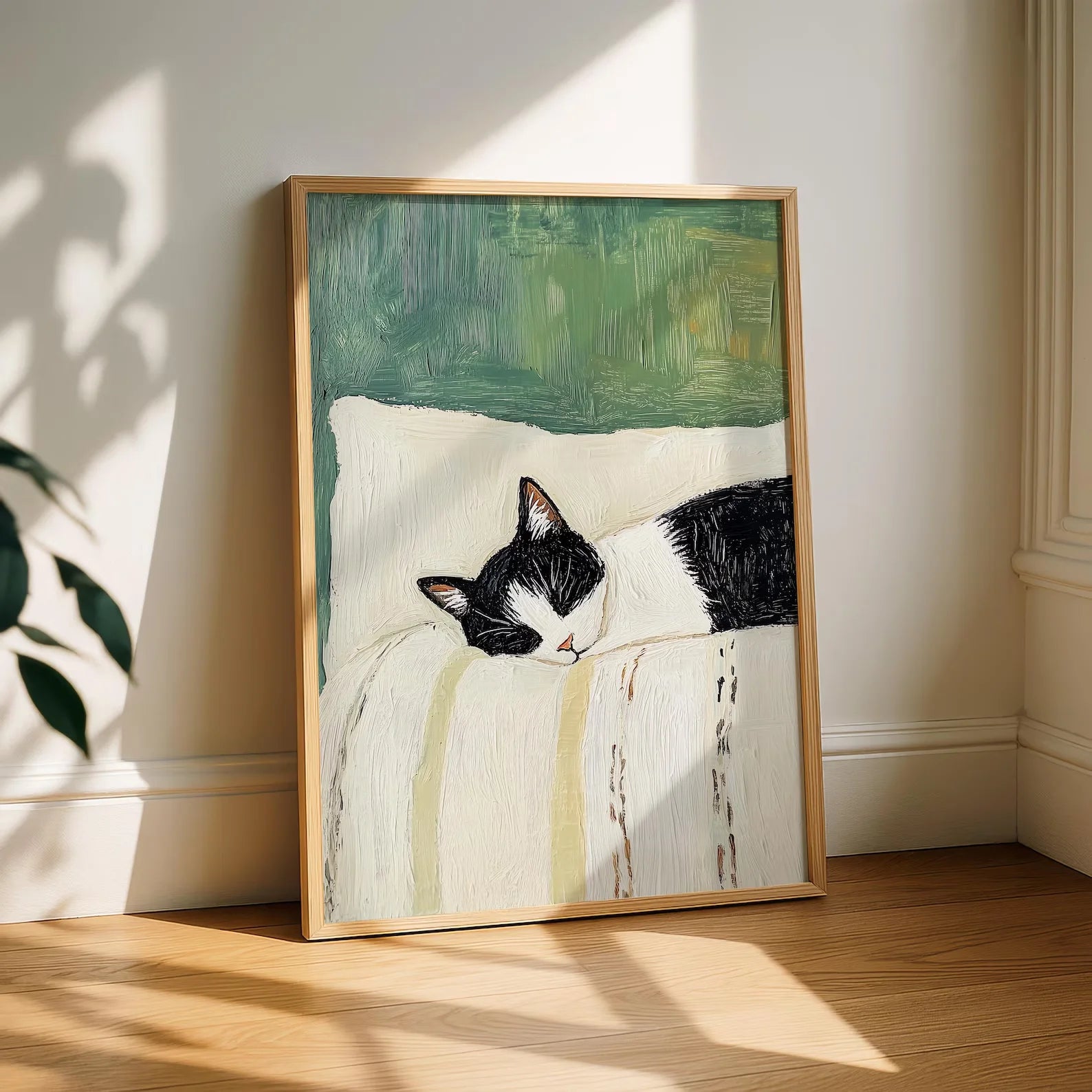 Framed artwork of a black and white cat on a pillow with a green background, leaning against a white wall.