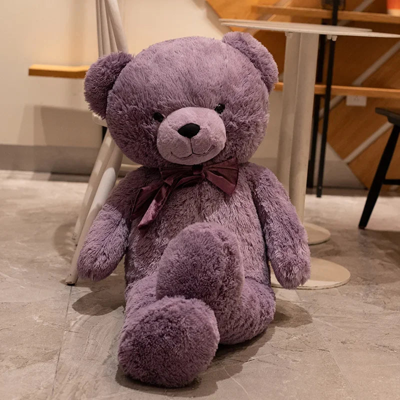 Purple teddy bear with a bow sitting on a tiled floor.