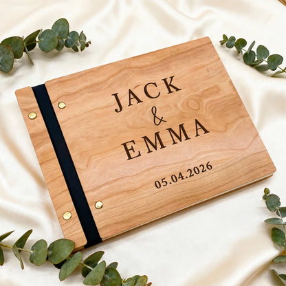 Wooden photo album with engraved names 'Jack & Emma' and date on a white fabric background with eucalyptus leaves.