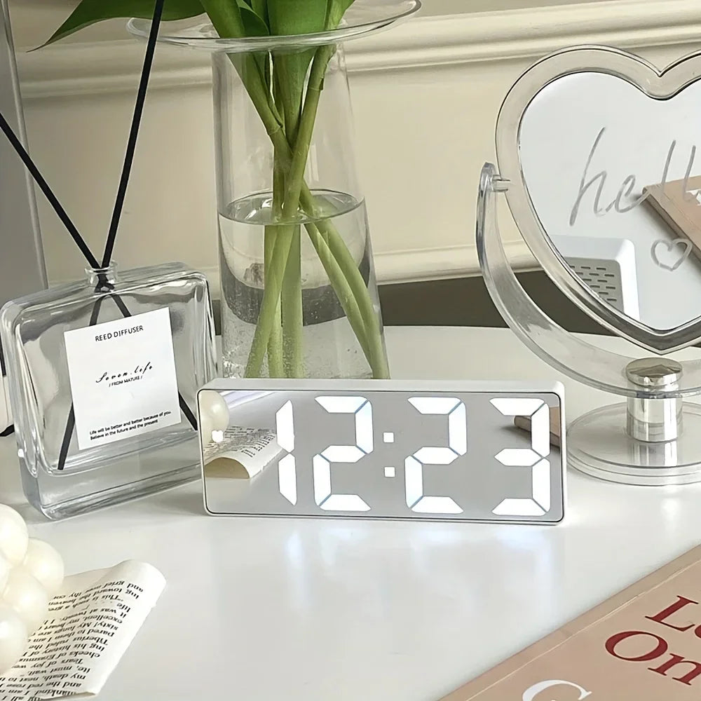 Digital clock on a table with decorative items including a diffuser, plant, and mirror.