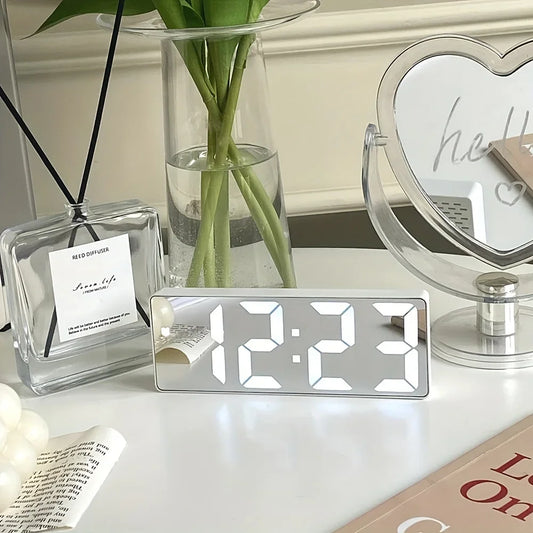 Digital clock on a table with decorative items including a diffuser, plant, and mirror.