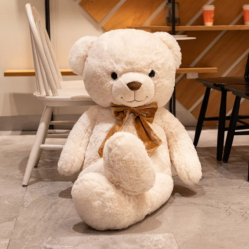 Large white teddy bear with a brown bow tie sitting on a wooden floor.