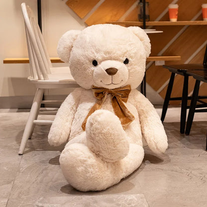 Large white teddy bear with a brown bow tie sitting on a wooden floor.
