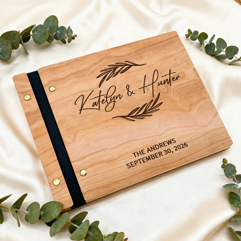 Wooden album with engraved names and date on a white fabric background with eucalyptus leaves.