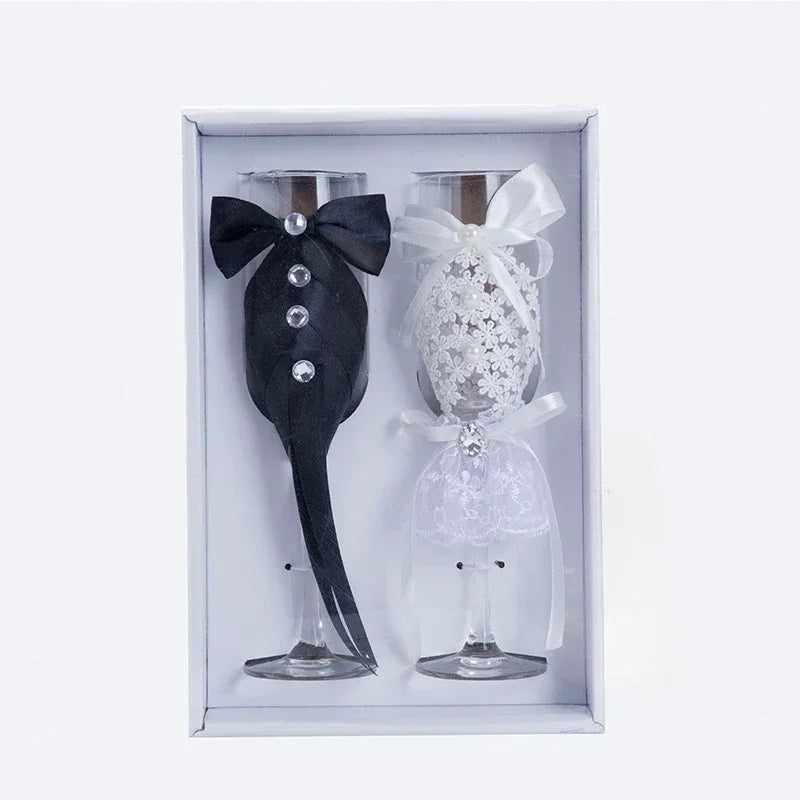 Two decorative wine glasses, one black with a bow tie and one white with lace design, in a box.