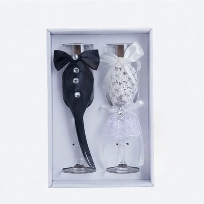 Two decorative wine glasses, one black with a bow tie and one white with lace design, in a box.