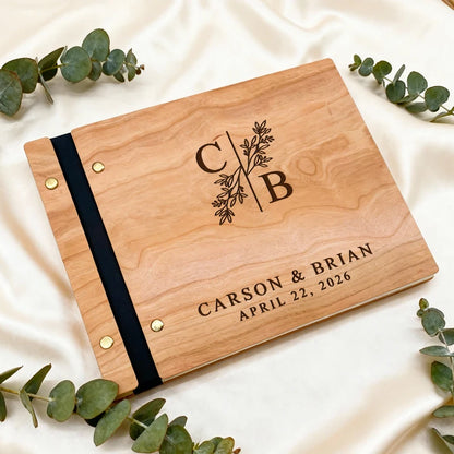 Wooden album with engraved initials and names on a white fabric background with eucalyptus leaves.