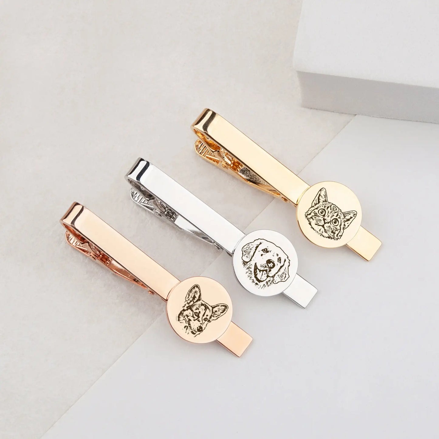 Three gold, silver, and rose gold tie clips with engraved dog designs on a light gray background.