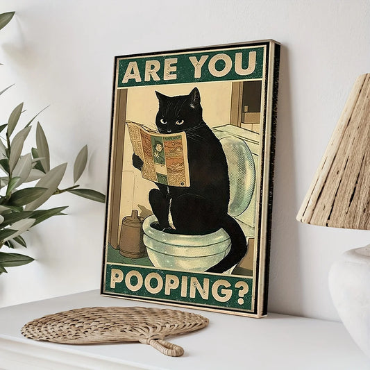 Framed poster with a black cat on a toilet and humorous text, placed on a surface with a plant and lamp in the background.