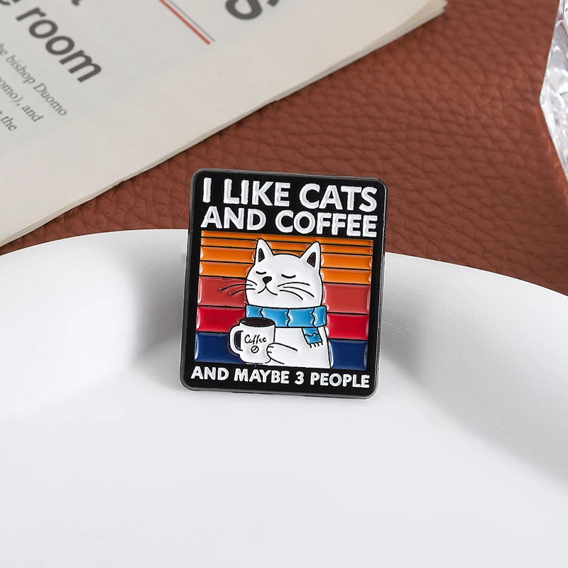 Enamel pin with a cat holding a coffee cup and text 'I like cats and coffee and maybe 3 people' on a white surface.