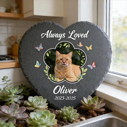 Heart-shaped slate plaque with a cat's photo and 'Always Loved Oliver 2023-2025' text, surrounded by plants.